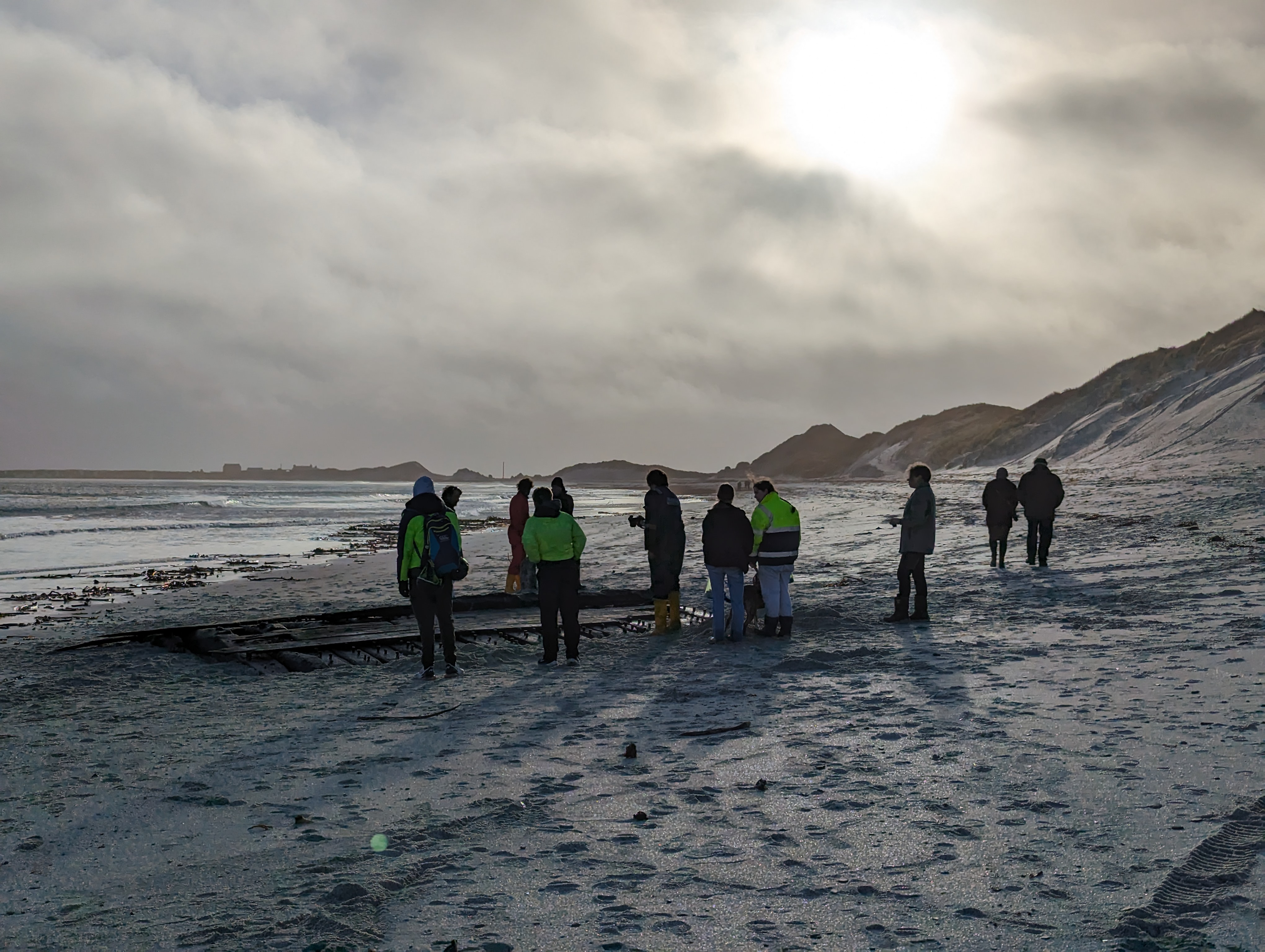 On a cold beach, a group of cold people look at a large section of oak wreckage rising from the sand